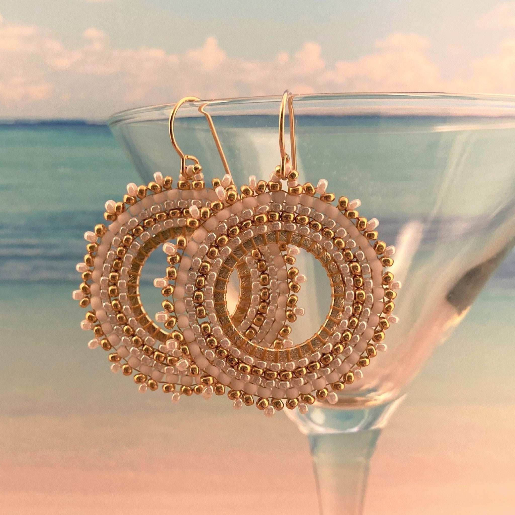 Petite White beaded hoops with gold beads around brushed gold hoops Beaded By The Beach displayed on a martini glass near a beach